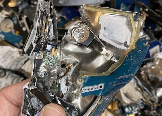 A person holding a piece of a hard drive in front of a pile of scrap metal.